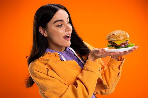 amazed woman in stylish jacket looking at tasty burger with cheese and meat isolated on orange
