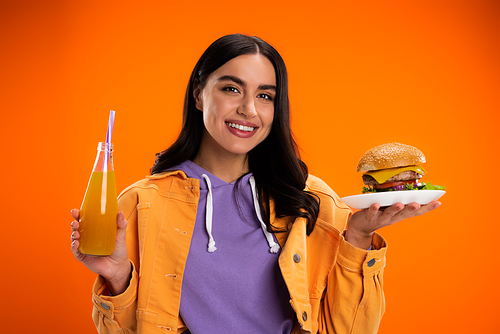 happy woman with delicious burger and bottle of fresh lemonade looking at camera isolated on orange