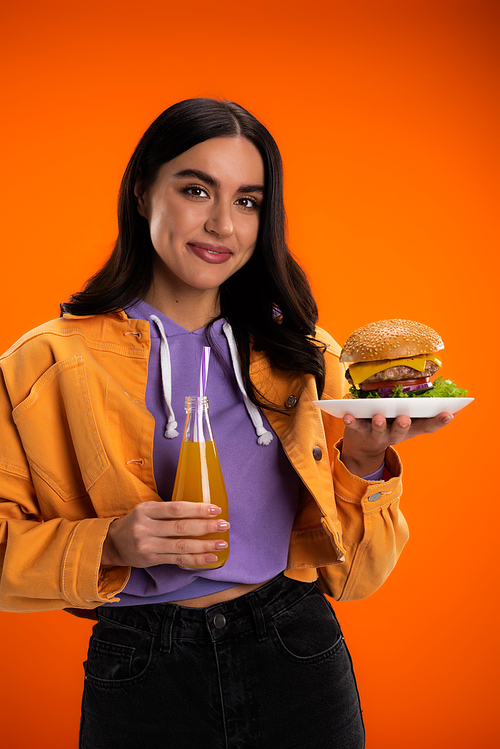 stylish woman with tasty burger and fresh lemonade smiling at the camera isolated on orange