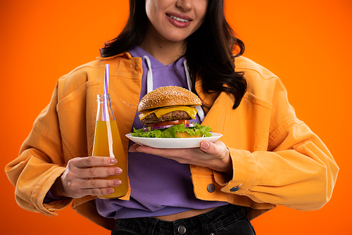 partial view of smiling woman with tasty burger and fresh lemonade isolated on orange