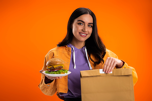 happy brunette woman holding paper bag and delicious burger isolated on orange
