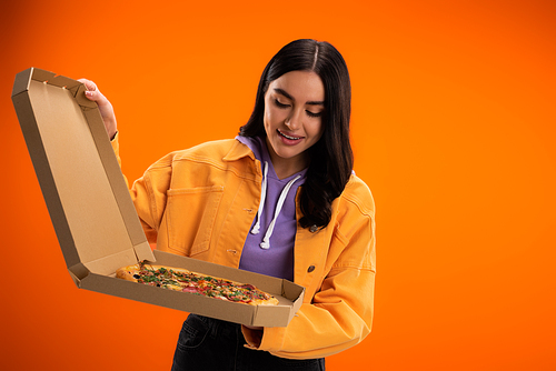 pleased woman in trendy jacket looking at tasty pizza in cardboard box isolated on orange