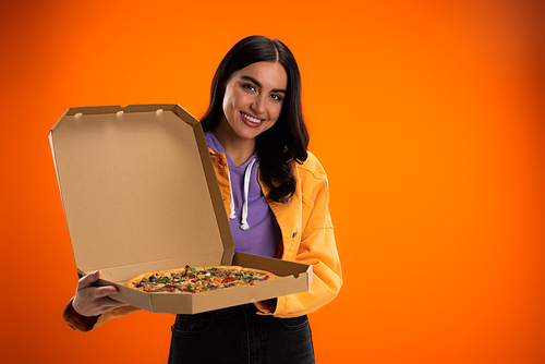 joyful woman looking at camera while holding carton box with tasty pizza isolated on orange
