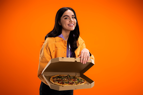 smiling brunette woman showing tasty pizza in carton box while looking at camera isolated on orange