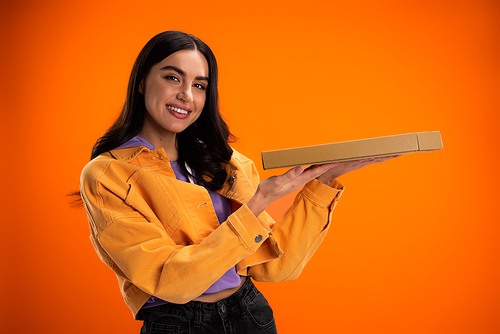 cheerful woman in trendy jacket holding pizza box and looking at camera isolated on orange