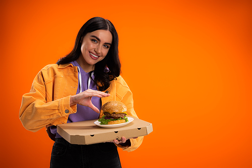 brunette woman with delicious burger and pizza box smiling at camera isolated on orange