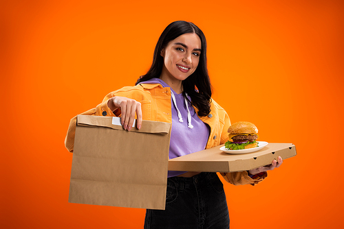 smiling woman holding tasty burger and paper bag while looking at camera isolated on orange