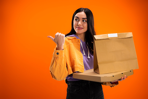 young woman with paper bag and pizza box looking away and pointing with thumb isolated on orange