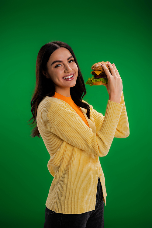 cheerful brunette woman looking at camera while holding tasty burger isolated on green