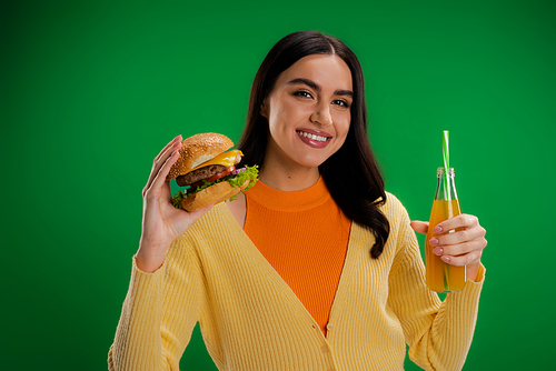 happy brunette woman holding delicious burger and bottle of fresh juice isolated on green