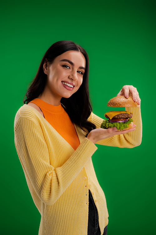 young brunette woman showing delicious burger while smiling at camera isolated on green