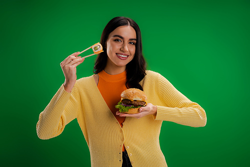 happy young woman holding burger and chopsticks with sushi roll isolated on green