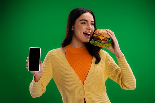 young woman with delicious burger showing smartphone with blank screen isolated on green