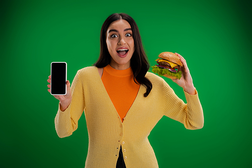 astonished woman holding tasty burger and mobile phone with blank screen isolated on green