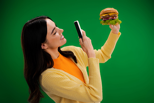 smiling woman taking photo of delicious burger on smartphone isolated on green