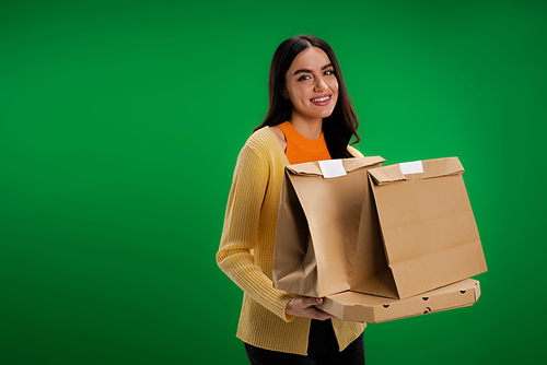 happy brunette woman holding paper bags and pizza box while looking at camera isolated on green