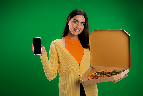 smiling woman with box of delicious pizza showing smartphone with blank screen isolated on green