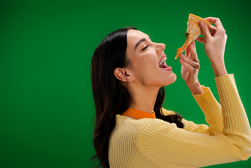 side view of young and hungry woman eating piece of pizza isolated on green