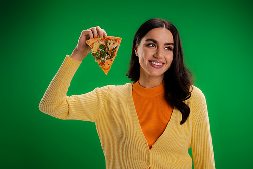 cheerful woman in orange turtleneck and yellow jumper holding piece of pizza and looking away isolated on green