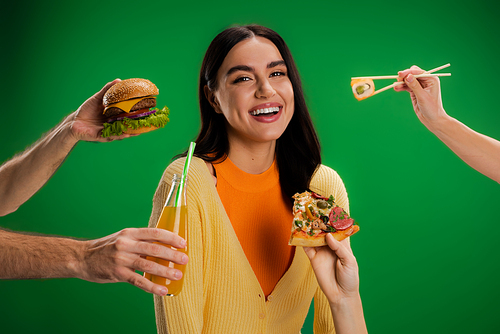 happy young woman looking at camera near people proposing different food isolated on green