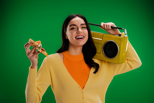 joyful woman with piece of delicious pizza and boombox looking at camera isolated on green
