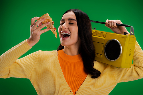 hungry woman with boombox holding piece of pizza near open mouth isolated on green