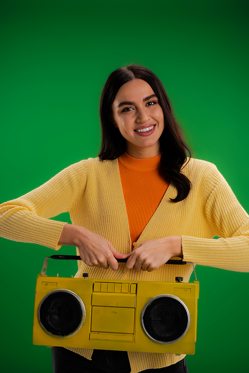 brunette woman in yellow jumper and orange turtleneck holding boombox and looking at camera isolated on green