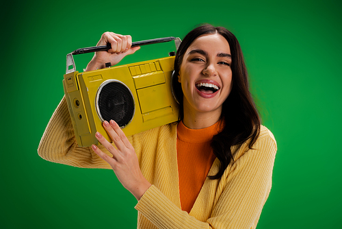 excited brunette woman holding boombox and laughing at camera isolated on green