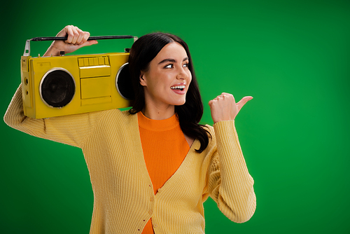 smiling woman with vintage boombox looking away and pointing with thumb isolated on green