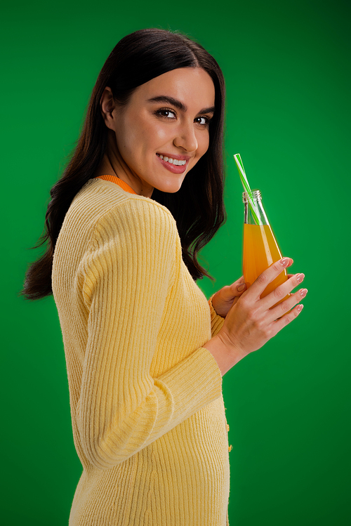 happy brunette woman looking at camera while standing with bottle of fresh juice isolated on green