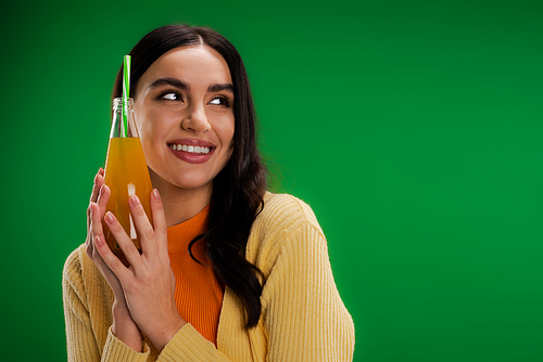 joyful brunette woman holding bottle of natural lemonade and looking away isolated on green