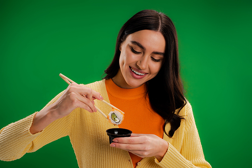smiling brunette woman holding sushi roll near sauce bowl isolated on green