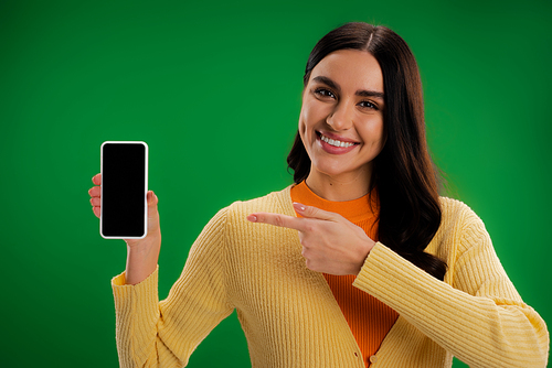 smiling brunette woman pointing at smartphone with blank screen isolated on green