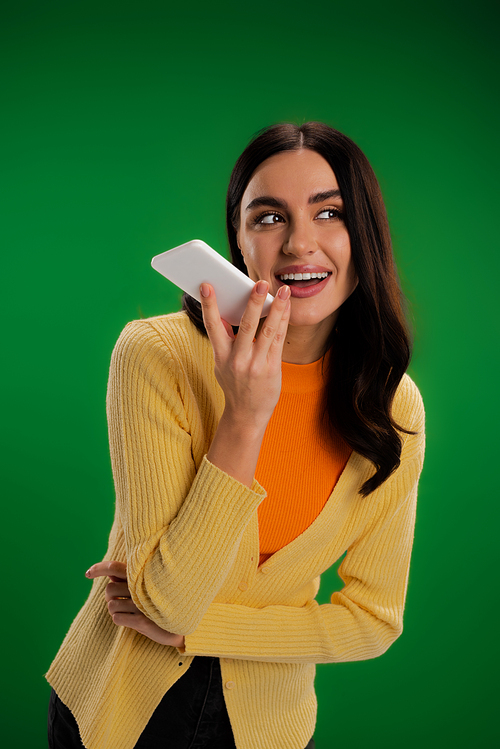cheerful brunette woman in orange turtleneck and yellow jumper recording voice message on smartphone isolated on green