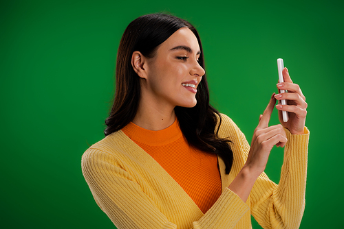 smiling brunette woman in yellow jumper and orange turtleneck using mobile phone isolated on green