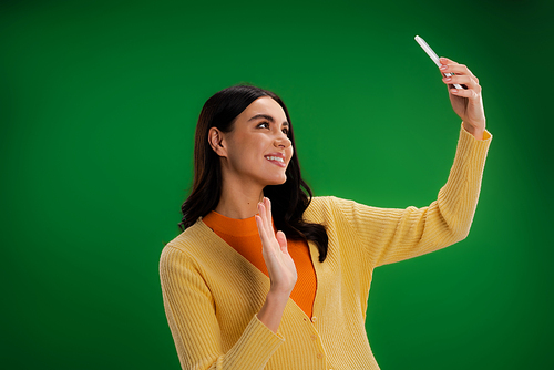 happy and stylish woman waving hand during video call on smartphone isolated on green