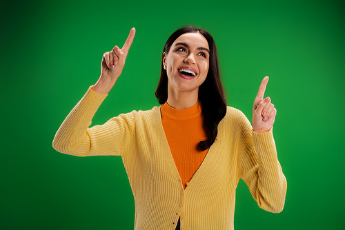 excited woman in yellow jumper looking up and pointing with fingers isolated on green