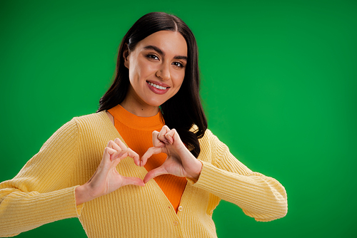 pretty brunette woman showing heart sign with hands and smiling at camera isolated on green