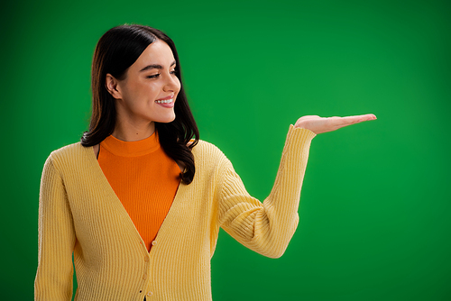smiling woman in orange turtleneck and yellow jumper pointing with hand isolated on green