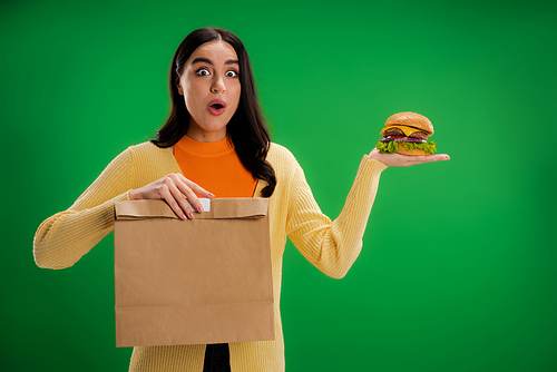 amazed woman with tasty burger and paper bag looking at camera isolated on green