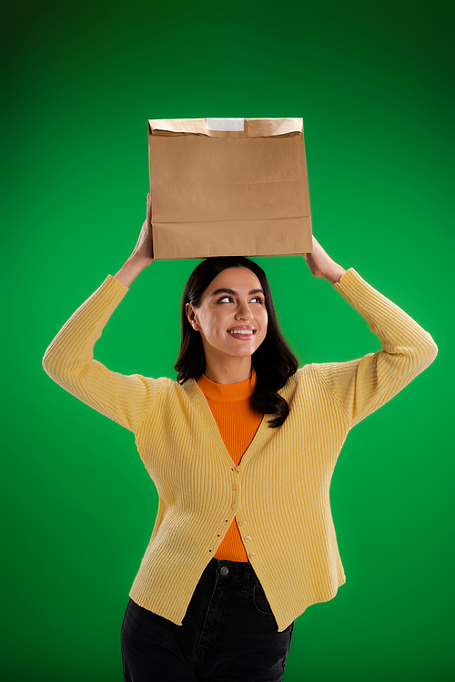 cheerful young woman holding paper bag above head isolated on green