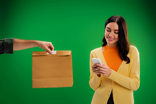 smiling brunette woman using smartphone near delivery man with paper bag isolated on green