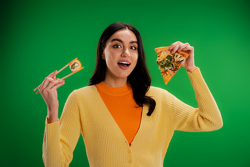 amazed woman looking at camera while holding tasty pizza and fresh sushi roll isolated on green