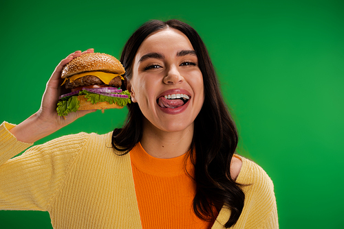 brunette woman holding tasty burger and sticking out tongue isolated on green