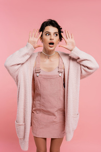 shocked woman in strap dress and warm cardigan standing with open mouth and showing wow gesture isolated on pink