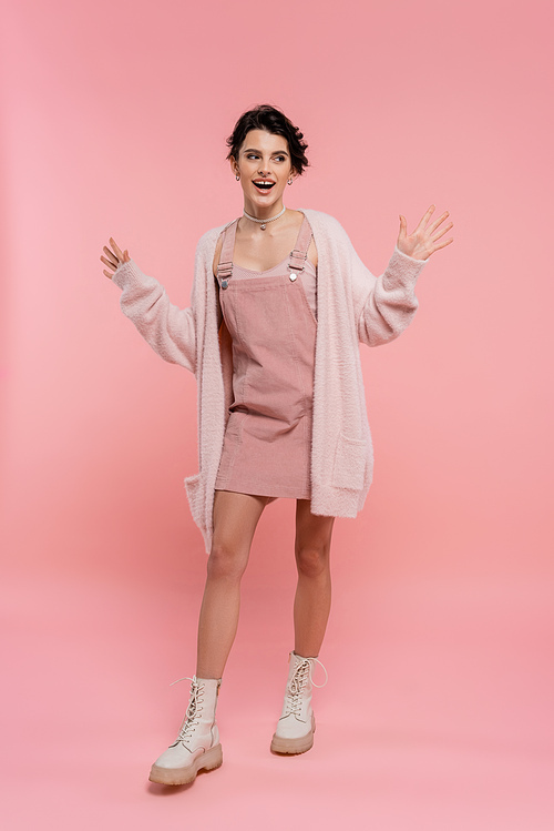 full length of excited woman in strap dress and fluffy cardigan showing wow gesture and looking away on pink background