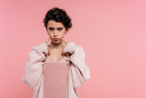 offended woman in fluffy cardigan touching puffed cheeks while looking at camera isolated on pink