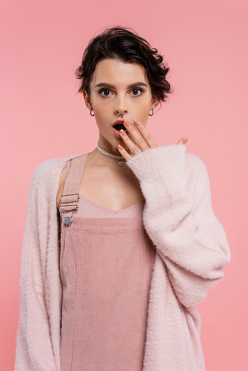 shocked woman in strap dress and warm cardigan covering open mouth with hand isolated on pink