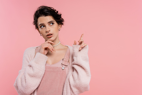 thoughtful brunette woman touching chin and pointing with finger isolated on pink