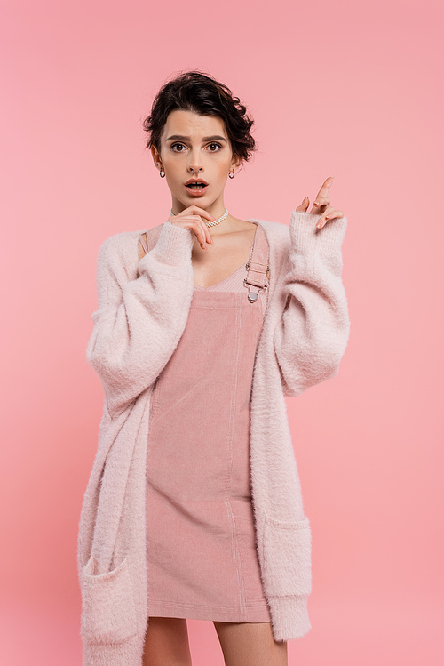 astonished woman in cozy cardigan touching chin and pointing with finger isolated on pink
